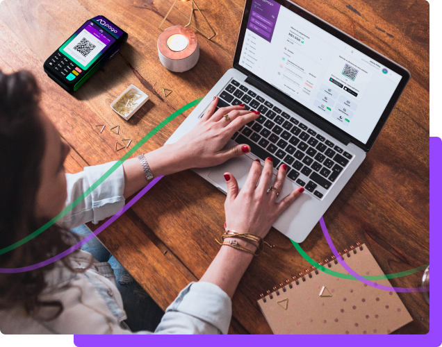 Mulher branca vestindo jeans azul claro, relgócio e pulseira d coura com unhas pintadas de vermelho digitando em um computador MacBook Pro com o internet banking da AqBank aberto. O computador está em cima de uma mesa de madeira junto de um caderno, uma máquina de cartão de crédito da AqPago e alguns grampos de folha de papel